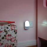 AVSL LYYT LED night light with PIR motion sensor illuminated in a white moulded housing, plugged into a UK socket on a pink wall beside a floral storage box.