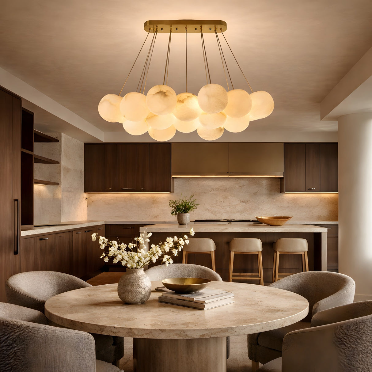 A modern kitchen with dark wood cabinets, a round stone dining table and four gray chairs is illuminated by the Noctis 9 Light Small Floating Globes Alabaster Chandelier in brushed brass, softly glowing overhead.