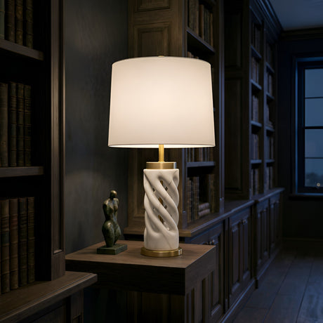 The Iverne 1 Light Twisted Carved Alabaster Table Lamp in Brass & Soft White sits on a wooden side table beside an abstract sculpture, framed by bookshelves in a dimly lit study.