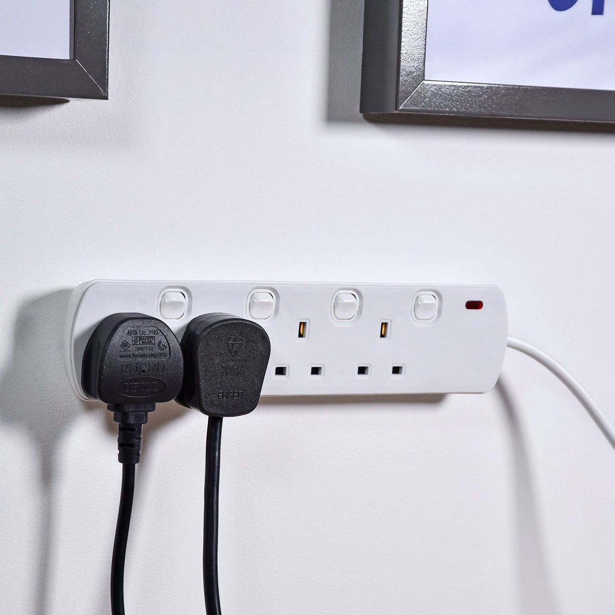 AVSL Mercury 4 gang switched extension lead in white mounted on a wall with two black UK plugs connected, below dark picture frames.