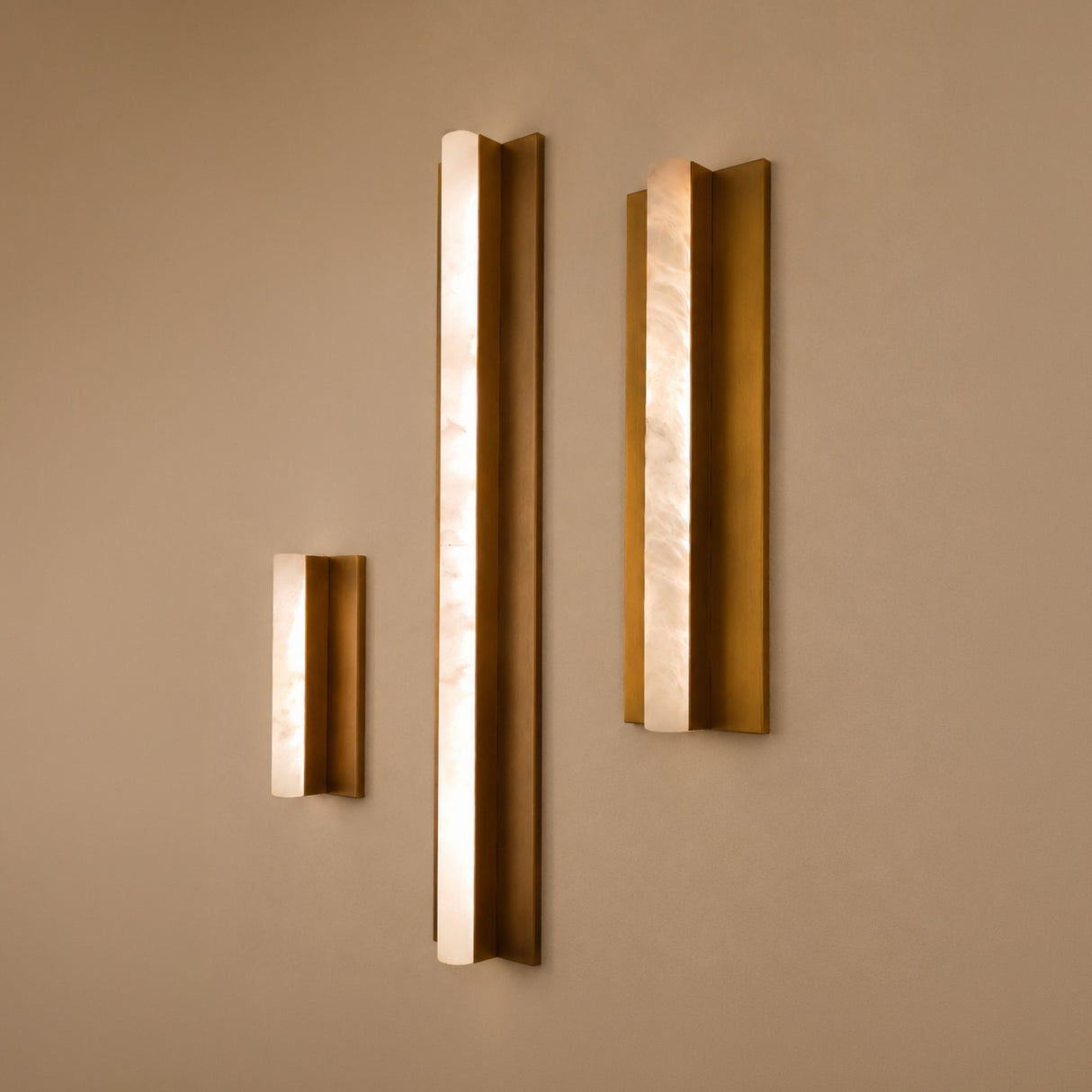Three Elvaris vertical wall lights in varying heights, featuring slender alabaster bars set within brushed brass backplates, mounted on a neutral wall.