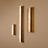 Three Elvaris vertical wall lights in varying heights, featuring slender alabaster bars set within brushed brass backplates, mounted on a neutral wall.