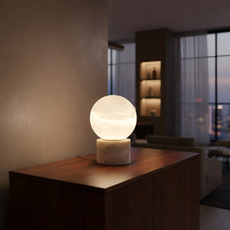 The Aurion LED Small Globe Alabaster Table Lamp - Soft White rests on a wooden cabinet in a modern living room with large windows and shelves, gently lighting the area at dusk.