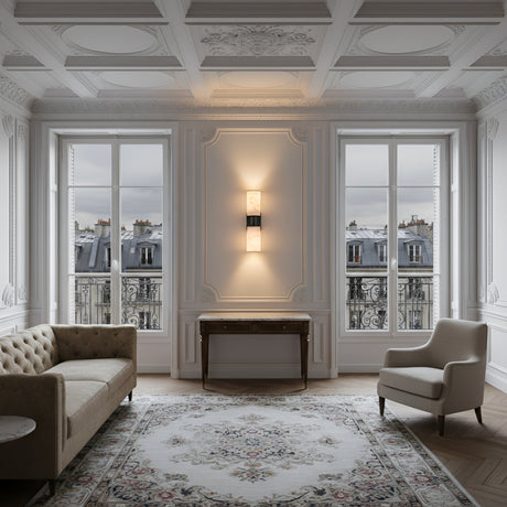 A bright, elegant living room with ornate white walls, large windows over Paris rooftops, a beige tufted sofa, armchair, wooden console table, patterned rug, and the Lumara 2 Light Twin Tube Alabaster Wall Light for a refined touch.
