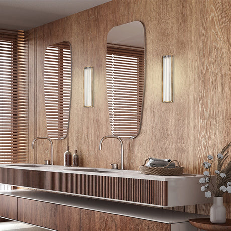 Kalvenne LED Glass Wall Light in satin bronze with ribbed glass shade, shown illuminated beside a mirror in a wood-panelled bathroom vanity setting.