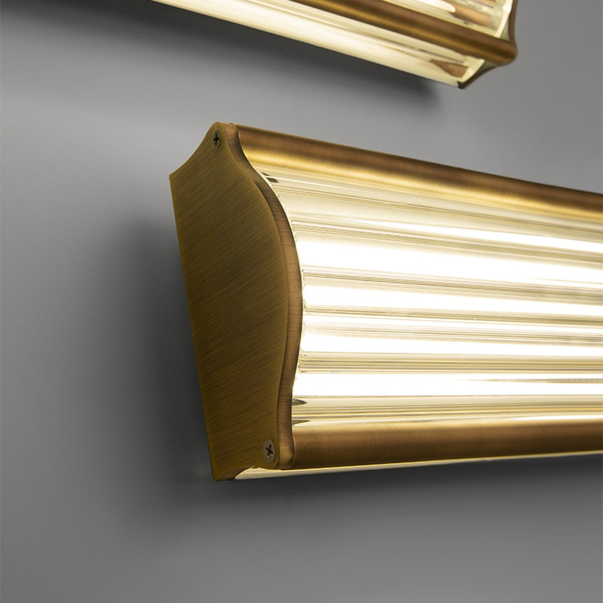 Kalvenne LED Glass Wall Light in satin bronze, close-up of ribbed glass shade and shaped bronze end cap with visible screw fixing, set against a grey wall.