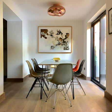 A modern dining room with a white table, six mismatched chairs, abstract wall art, the Lava Large LED Wall & Ceiling Light in copper, and large glass doors that fill the space with natural light.