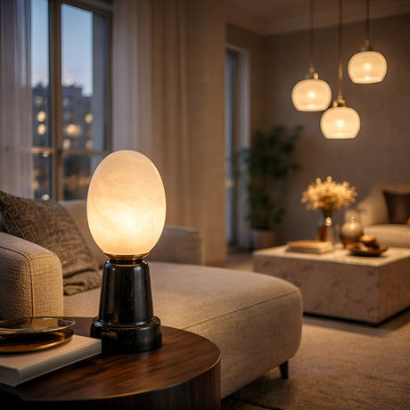 A cozy living room at dusk features the Ovolis 1 Light Oval Alabaster Table Lamp in soft white & black on a side table beside beige sofas, with a decorated coffee table and three pendant lights. Subtle tones recall natural Spanish stone warmth.