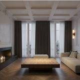 A modern, minimalistic living room with a beige sofa, wooden coffee table, fireplace on the left, large windows with sheer and dark curtains, herringbone floors, and the Lumara 2 Light Twin Tube Alabaster Wall Light in soft white & brushed brass.
