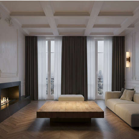 A modern, minimalistic living room with a beige sofa, wooden coffee table, fireplace on the left, large windows with sheer and dark curtains, herringbone floors, and the Lumara 2 Light Twin Tube Alabaster Wall Light in soft white & brushed brass.