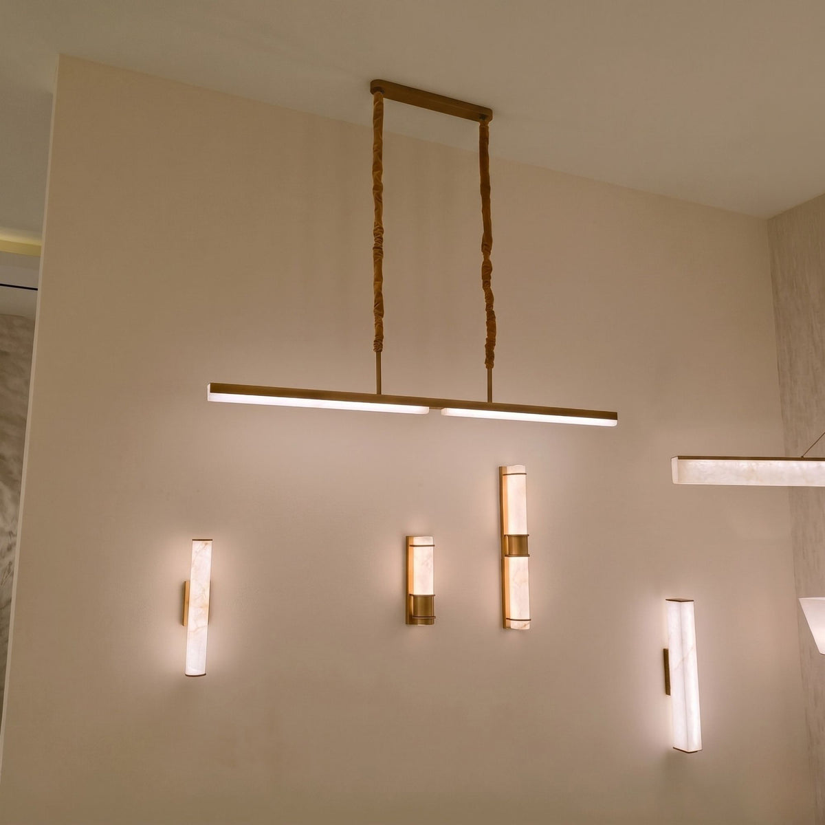 Elvaris LED Linear Alabaster Chandelier in brushed brass, shown suspended from a ceiling plate on twisted cords above a wall with matching alabaster wall lights.