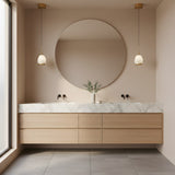 A modern bathroom with a large round mirror, light wood vanity with marble countertop, double sinks with black fixtures, and a vase of greenery. Natural light is enhanced by the Aurevia LED Drop Alabaster Single Pendant Light in brushed brass.