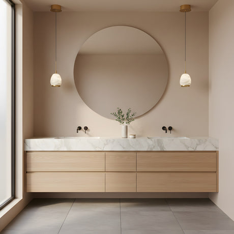 A modern bathroom with a large round mirror, light wood vanity with marble countertop, double sinks with black fixtures, and a vase of greenery. Natural light is enhanced by the Aurevia LED Drop Alabaster Single Pendant Light in brushed brass.