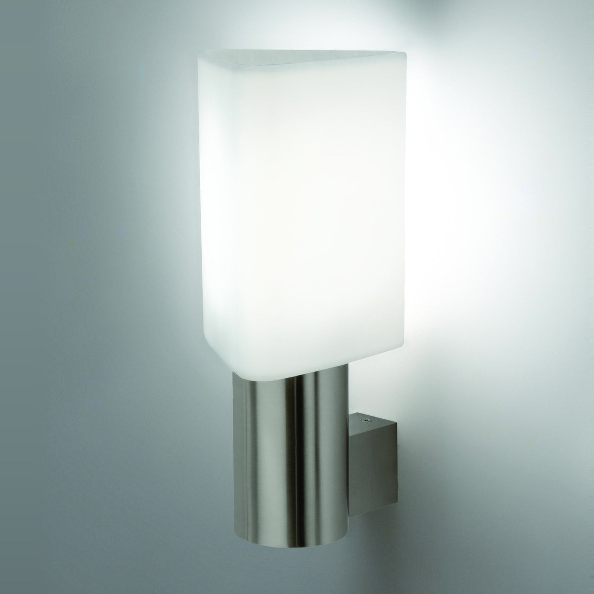 Tiny Triangle Wall Light in satin nickel, with a white triangular glass shade on a slim cylindrical column mounted via a square bracket, shown lit against a plain light grey wall.