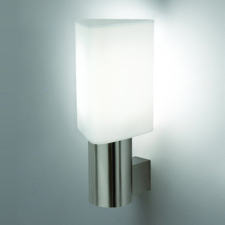 Tiny Triangle Wall Light in satin nickel, with a white triangular glass shade on a slim cylindrical column mounted via a square bracket, shown lit against a plain light grey wall.