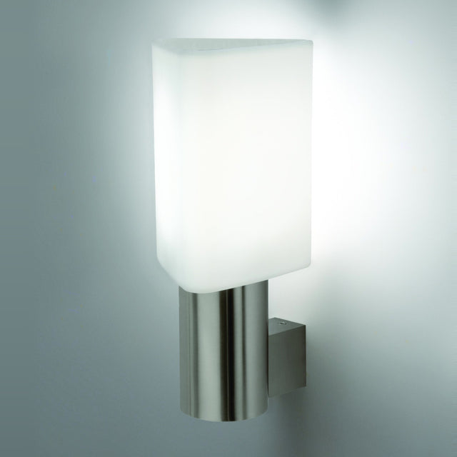 Tiny Triangle Wall Light in satin nickel, with a white triangular glass shade on a slim cylindrical column mounted via a square bracket, shown lit against a plain light grey wall.