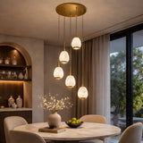 A modern dining room with a round table, beige chairs, a vase with branches, bowl of green apples, and the Aurevia 5 Light LED Drop Alabaster Cluster Pendant Light in Brushed Brass. Built-in shelves display pottery; large windows overlook greenery.