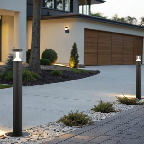 Tagus 100cm LED PIR lamp post in anthracite, a slim cylindrical outdoor bollard light with clear diffuser top, shown lining a modern house driveway at dusk.