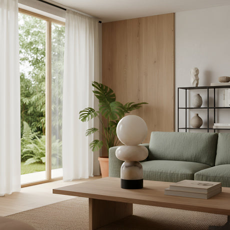 A modern living room features a light green sofa, a wooden coffee table with books and the Talmera 1 Light Small Sculptural Sphere Alabaster Table Lamp in soft white & black, a potted plant, a black shelving unit, and large windows with sheer white curtains.