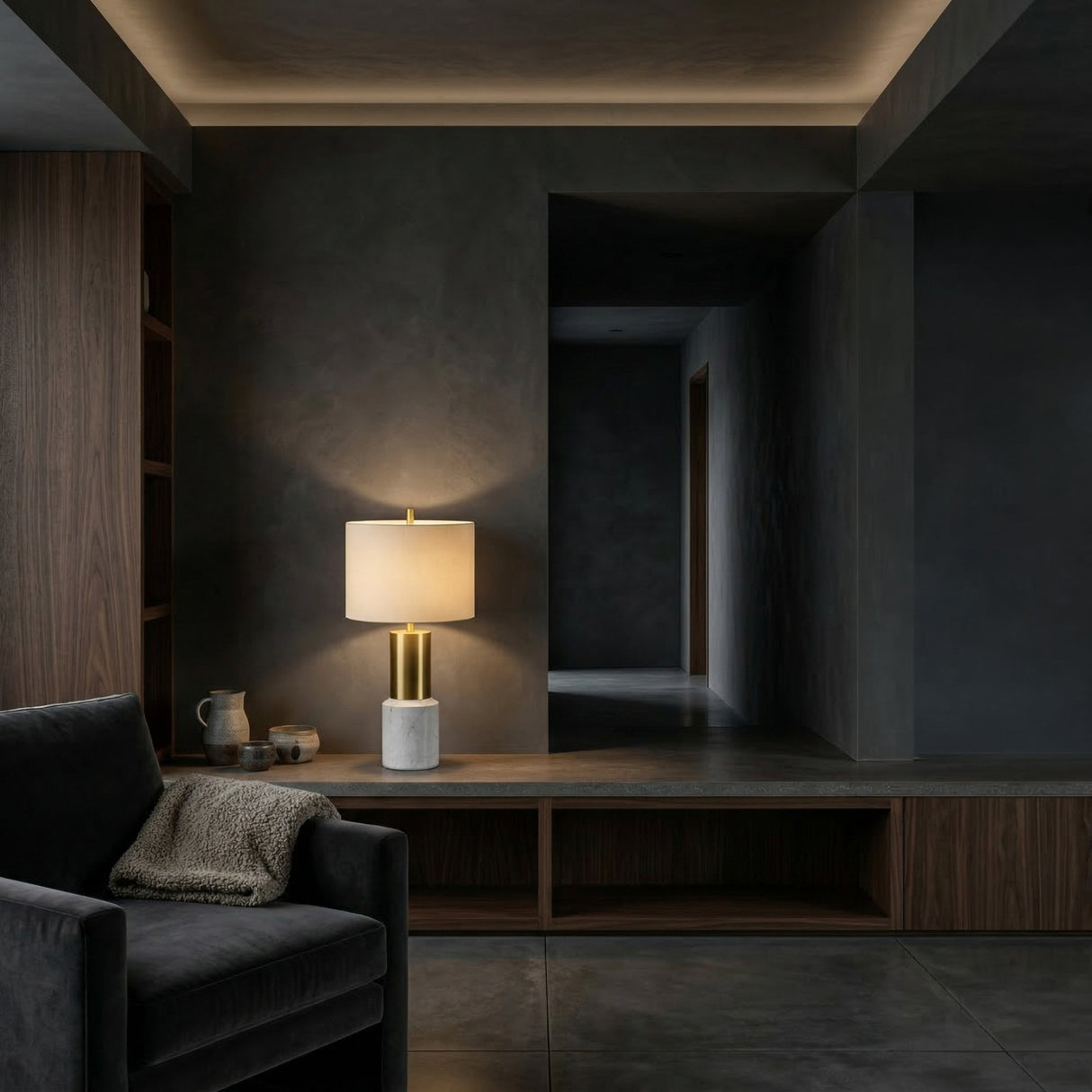 A modern, dimly lit living space with dark walls features a gray armchair with a knit blanket, a wooden built-in shelf, and the Lunaro 1 Light Marble Table Lamp – Brass & White casting a warm glow.