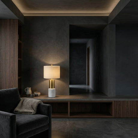 A modern, dimly lit living space with dark walls features a gray armchair with a knit blanket, a wooden built-in shelf, and the Lunaro 1 Light Marble Table Lamp – Brass & White casting a warm glow.
