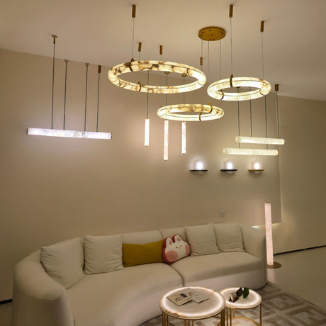 Aureon LED Triple Ring Alabaster Chandelier in brushed brass, shown with matching linear and tube pendants illuminated above a beige sofa in a living room setting.