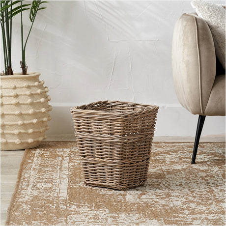Tobs Grey Kubu Rattan Waste Paper Bin, a tapered woven storage box, 35cm tall, shown on a neutral rug beside a plant pot and upholstered chair.