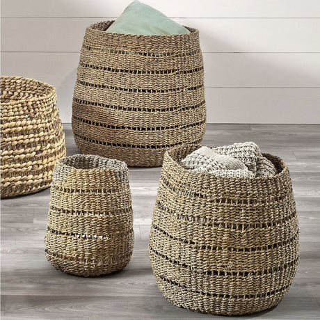 Set of 3 round woven seagrass storage baskets in natural finish, tapered shapes with visible stripe texture, shown holding folded throws on a wooden floor.