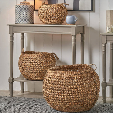 Set of 3 round water hyacinth handled storage baskets in natural wicker, shown in varying sizes on and beneath a grey console table in a living room setting.