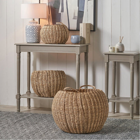 Set of 3 round natural seagrass and palm leaf storage baskets with woven handles, shown in a hallway on a console table, under-table shelf, and floor.