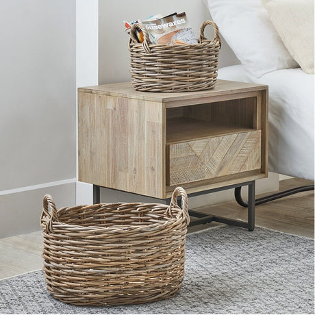 Set of 2 grey Kubu oval baskets with woven rattan texture and looped handles, shown beside a wooden bedside table in a bedroom setting.
