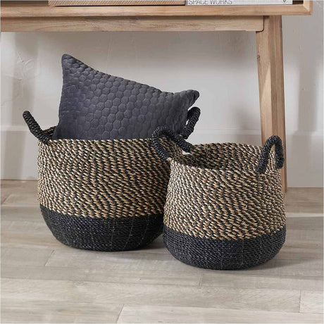 Set of 2 seagrass handled baskets with natural woven bodies and black bases, shown in a room holding a grey quilted cushion beside a wooden bench.