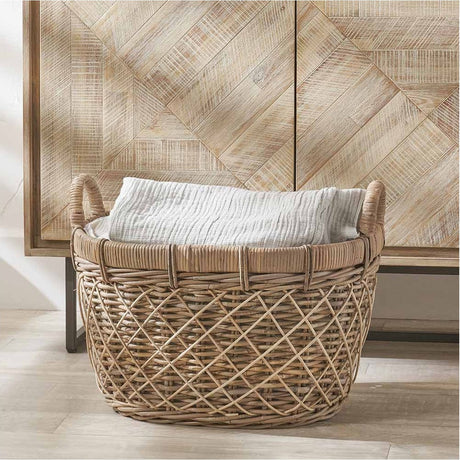 Brown rattan oval laundry basket with woven diamond pattern and two side handles, holding folded linen, shown beside a wood-panelled cabinet.