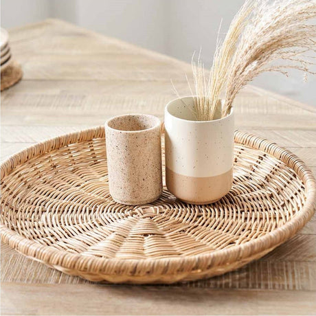 Rattan Basket Tray Tobs, 43cm round woven tray in natural finish, styled on a wooden table with a speckled ceramic tumbler and vase holding dried grasses.