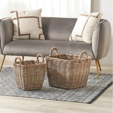 Set of 2 grey Kubu rattan storage baskets with woven handles, shown in graduated sizes on a rug beside a grey sofa with patterned cushions.