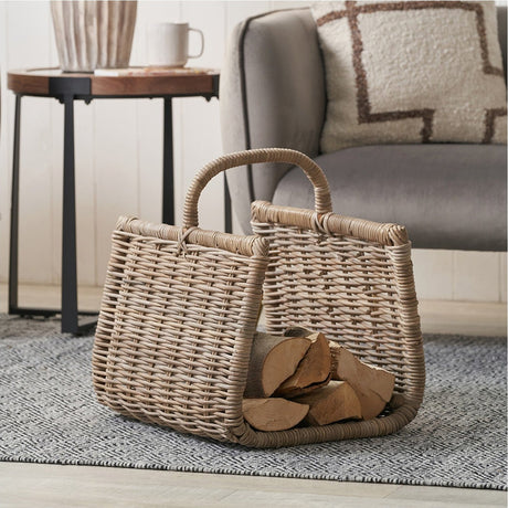 Grey Kubu Trug Basket - 51cm, a woven rattan storage basket with arched handle, shown holding logs beside a sofa and side table in a living room setting.