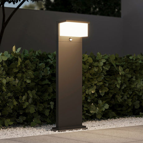 Nestos 80cm LED PIR lamp post in anthracite, a slim rectangular aluminium post with a rectangular illuminated diffuser head and motion sensor, shown lit at night beside a garden hedge.