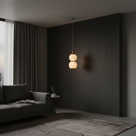 A modern, minimal living room with a dark textured accent wall, gray sofa, round side table holding a cup and book, sheer curtains, and the Lumina 2 Light Orb Alabaster Single Pendant Light in soft white & brass hanging from the ceiling.