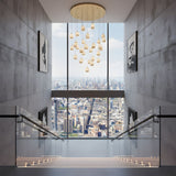 Niori Aurevia 31 Light LED alabaster chandelier in brushed brass, cascading pebble-shaped drops above a concrete stairwell with city skyline views.