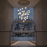 Niori Aurevia 31 Light LED alabaster chandelier in brushed brass, cascading pebble-shaped drops above a staircase with city skyline views.
