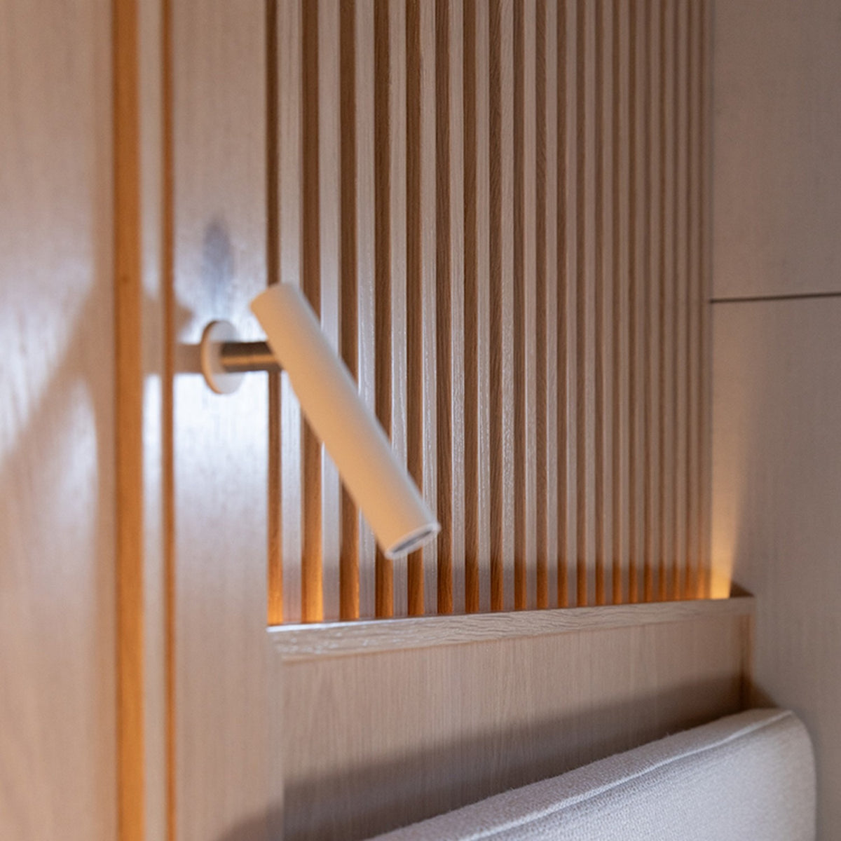 Ivelline LED wall light with reading light in white, slim cylindrical head on a short arm with round wall rosette, mounted on ribbed wood panelling in a bedroom setting.