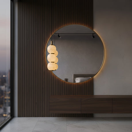 A modern, minimal interior features a large round backlit mirror on a wood-paneled wall. Reflected in the mirror is the Lumina 3 Light Orb Alabaster Single Pendant Light in Soft White & Brass. Marble floors and a city skyline are visible to the left.