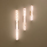 Montavo LED alabaster tube wall lights, 800mm cylinders in natural stone with brushed brass caps and backplates, mounted at varying heights, glowing warm white against a neutral wall.