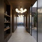 Modern hallway with dark wood walls, built-in shelves holding vases and plants, and the Ivy 9 Light Chandelier in brass overhead. Large windows at the end reveal a city view, while minimalist decor and sleek design complete the space.