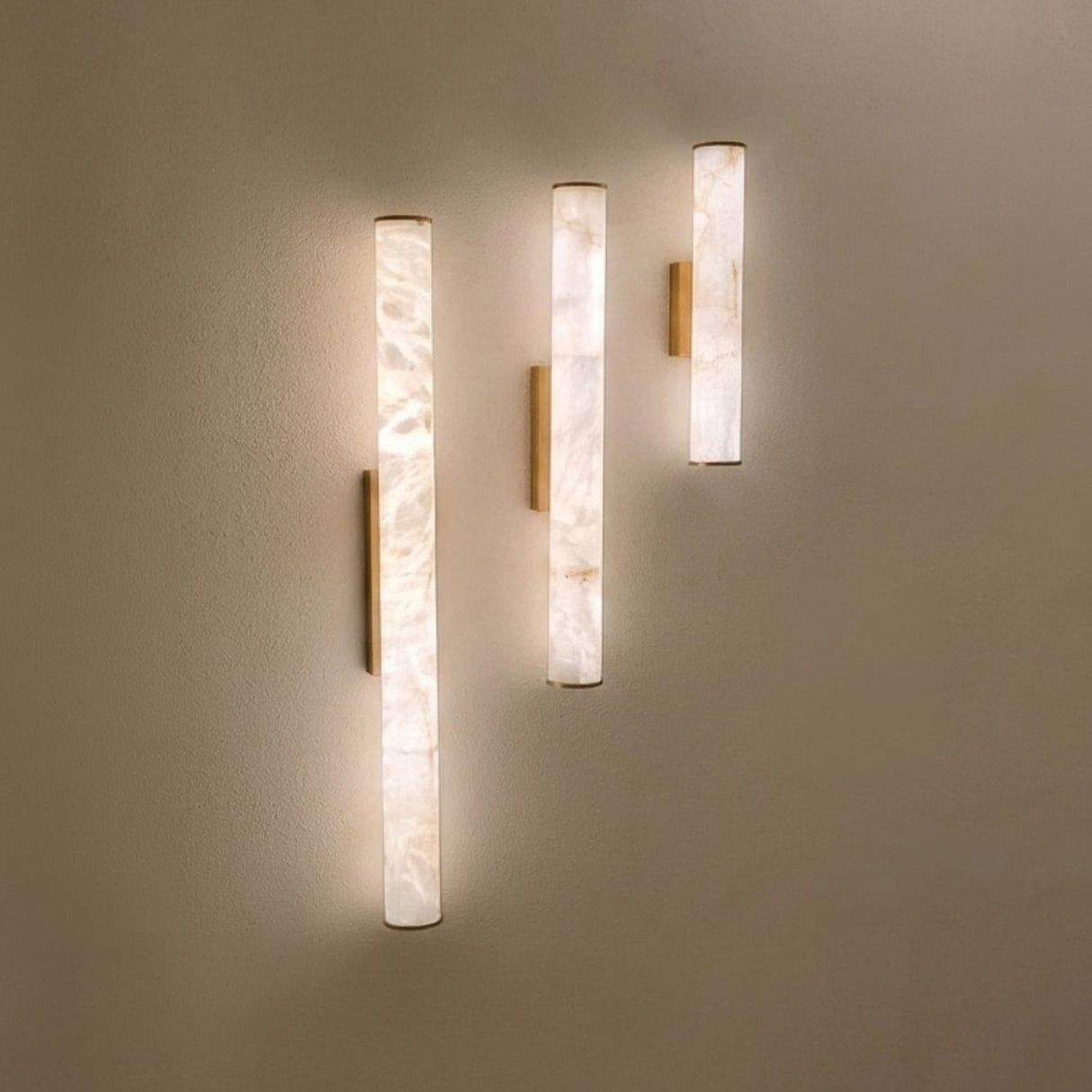 Montavo LED alabaster tube wall lights in three lengths with brushed brass caps and backplates, mounted on a plain wall showing warm illuminated glow.