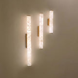 Montavo LED alabaster tube wall lights in three lengths with brushed brass caps and backplates, mounted on a plain wall showing warm illuminated glow.