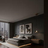 A modern bedroom with a large bed, abstract art above the headboard, floating nightstands, and an Aurevia LED Disc Alabaster Single Pendant Light in brushed brass casting soft light. Floor-to-ceiling windows reveal a city view with ambient under-bed lighting.