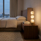 A modern bedroom with a neatly made bed, beige linens, and a padded headboard. A wooden nightstand holds the Orbelya 1 Light Stacked Alabaster Table Lamp in Soft White & Brass, plus books, beside a large window framing a dusk city view.