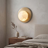 Oria Alabaster Globe Wall Light in brushed brass, a round backplate with a glowing alabaster sphere, shown lit above a bedside table in a bedroom.