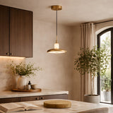 A modern kitchen with wood cabinets, marble countertop, potted plants, an open book, a round wooden board, and natural light streaming in through a large window with beige curtains features an Aurevia LED Disc Alabaster Single Pendant Light in brushed brass.